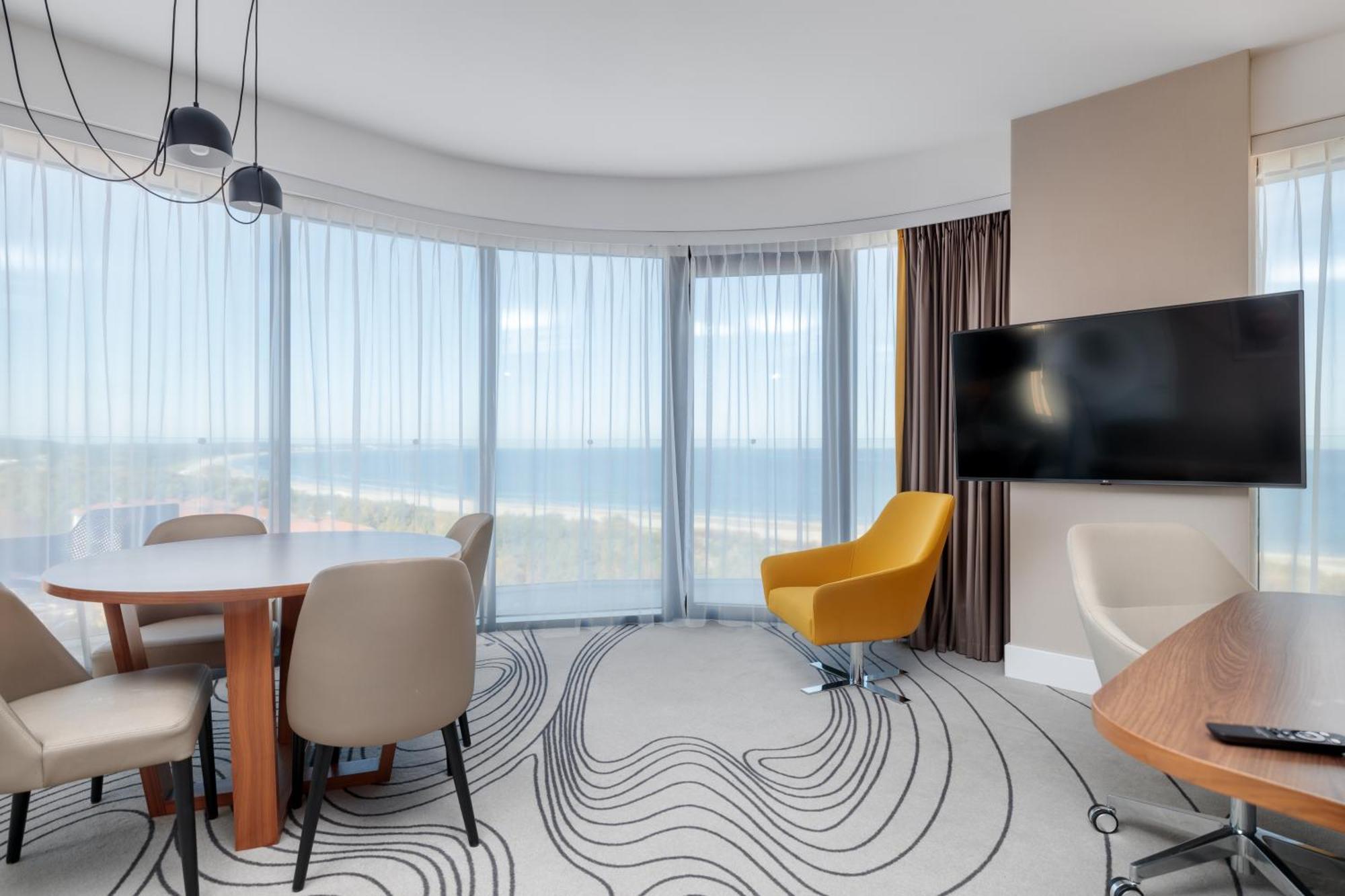 Radisson Blu Resort Świnoujście Exterior foto The image shows a modern hotel room or suite with a bright, airy atmosphere. There are large windows that allow natural light to fill the space, providing a view of the ocean. The decor includes a round dining table with several chairs, and a stylish