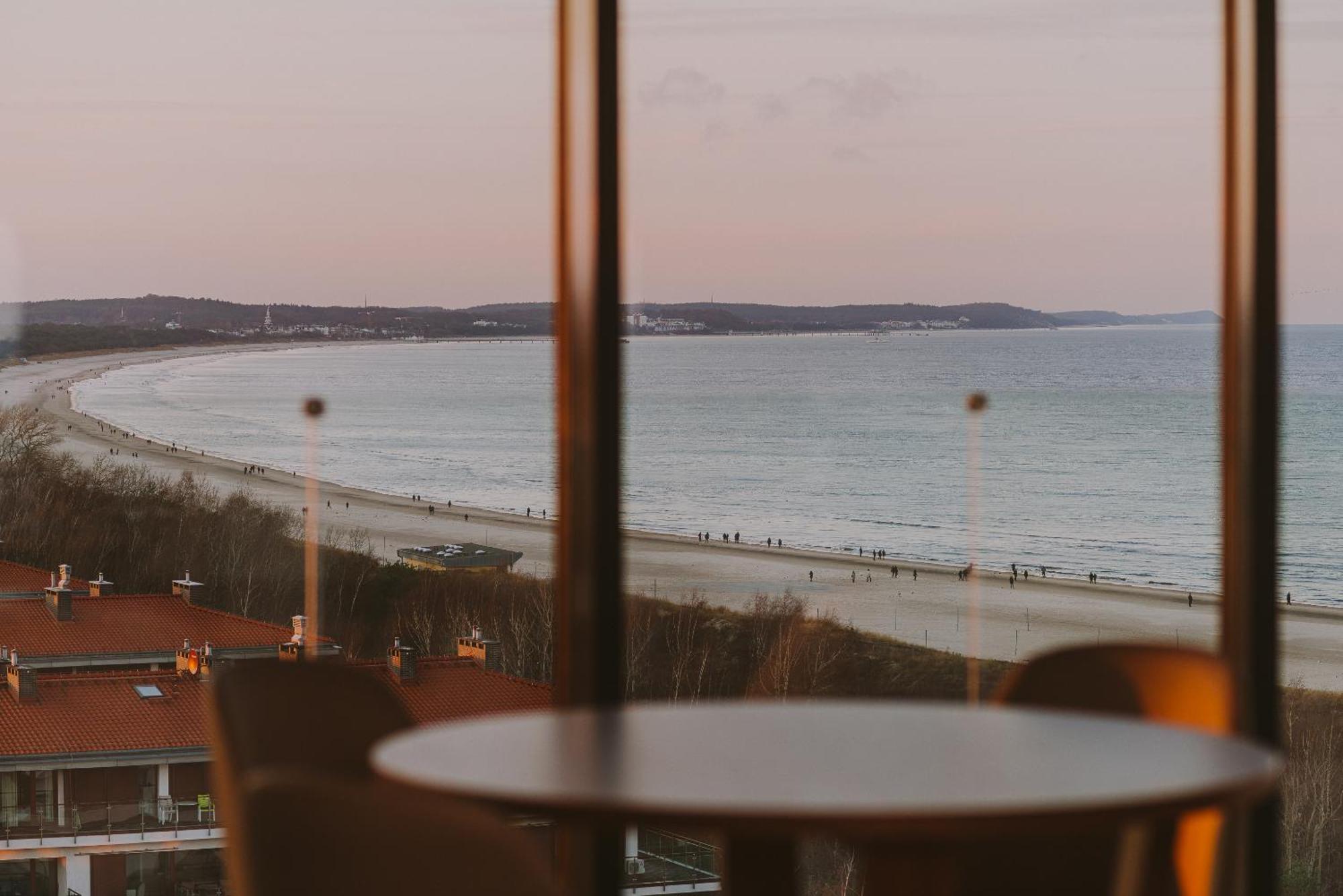 Radisson Blu Resort Świnoujście Exterior foto The photo shows a picturesque view of a beach scene from indoors. In the foreground, there is a round table with some chairs around it, suggesting a cozy interior space. Through a large window, you can see the beach with gentle waves hitting the shor