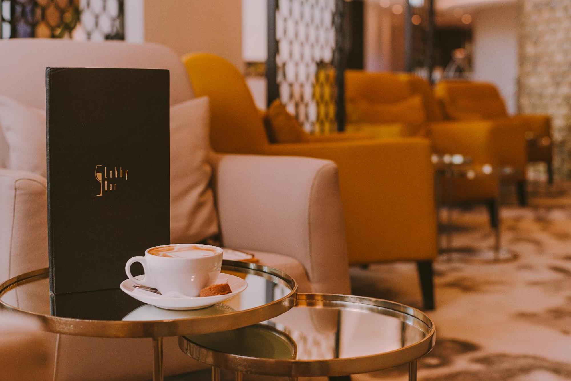 Radisson Blu Resort Świnoujście Exterior foto The photo shows a cozy indoor setting with a stylish, modern atmosphere. In the foreground, there is a small round glass table with a white coffee cup and a saucer, alongside a small biscuit or cookie. Next to the coffee, there is a black menu with e