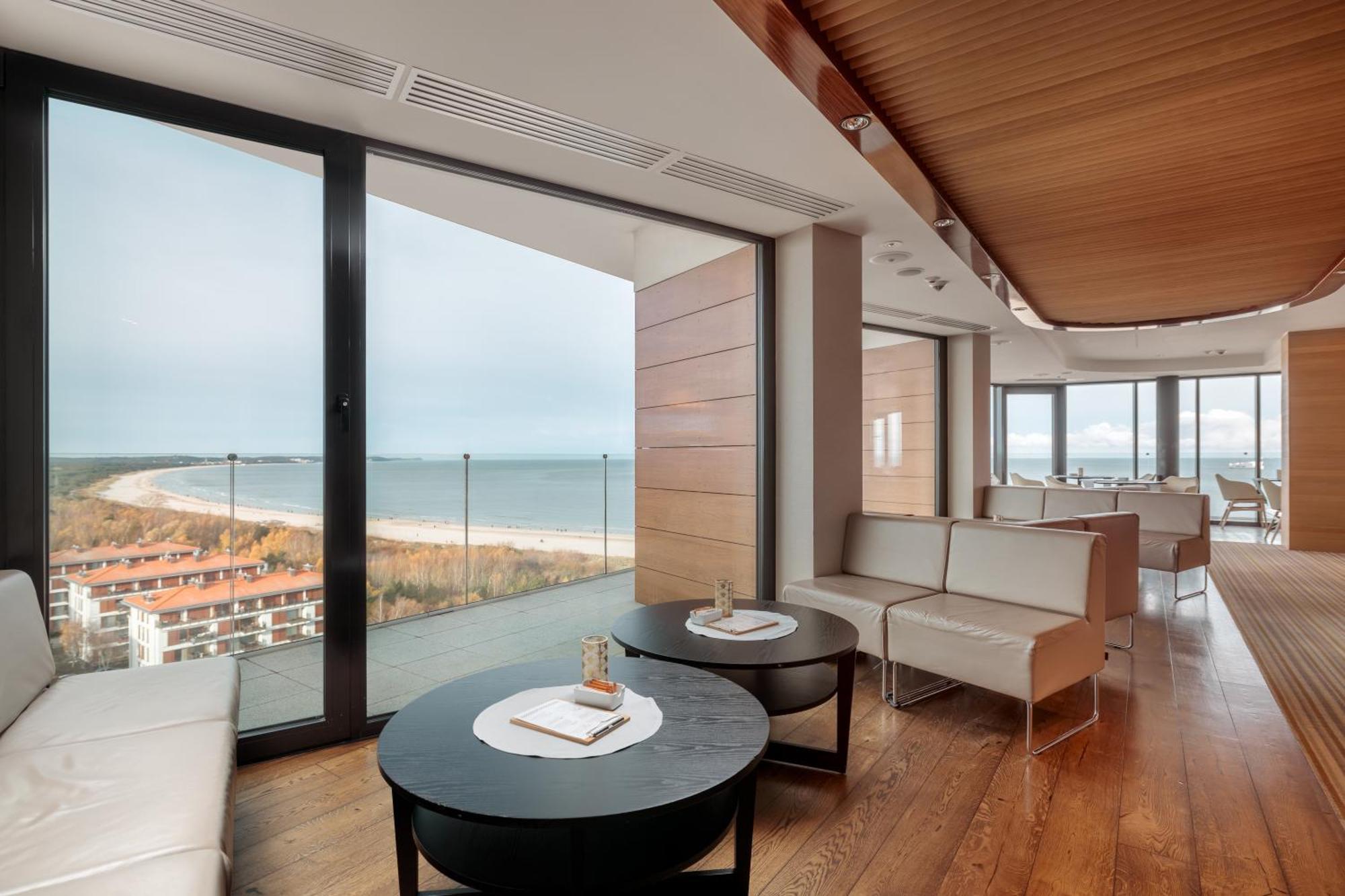 Radisson Blu Resort Świnoujście Exterior foto The photo shows a modern, elegantly designed interior space featuring large windows that offer a panoramic view of the ocean and coastline. The flooring is wooden, providing a warm atmosphere. There are two round black tables in the foreground with m
