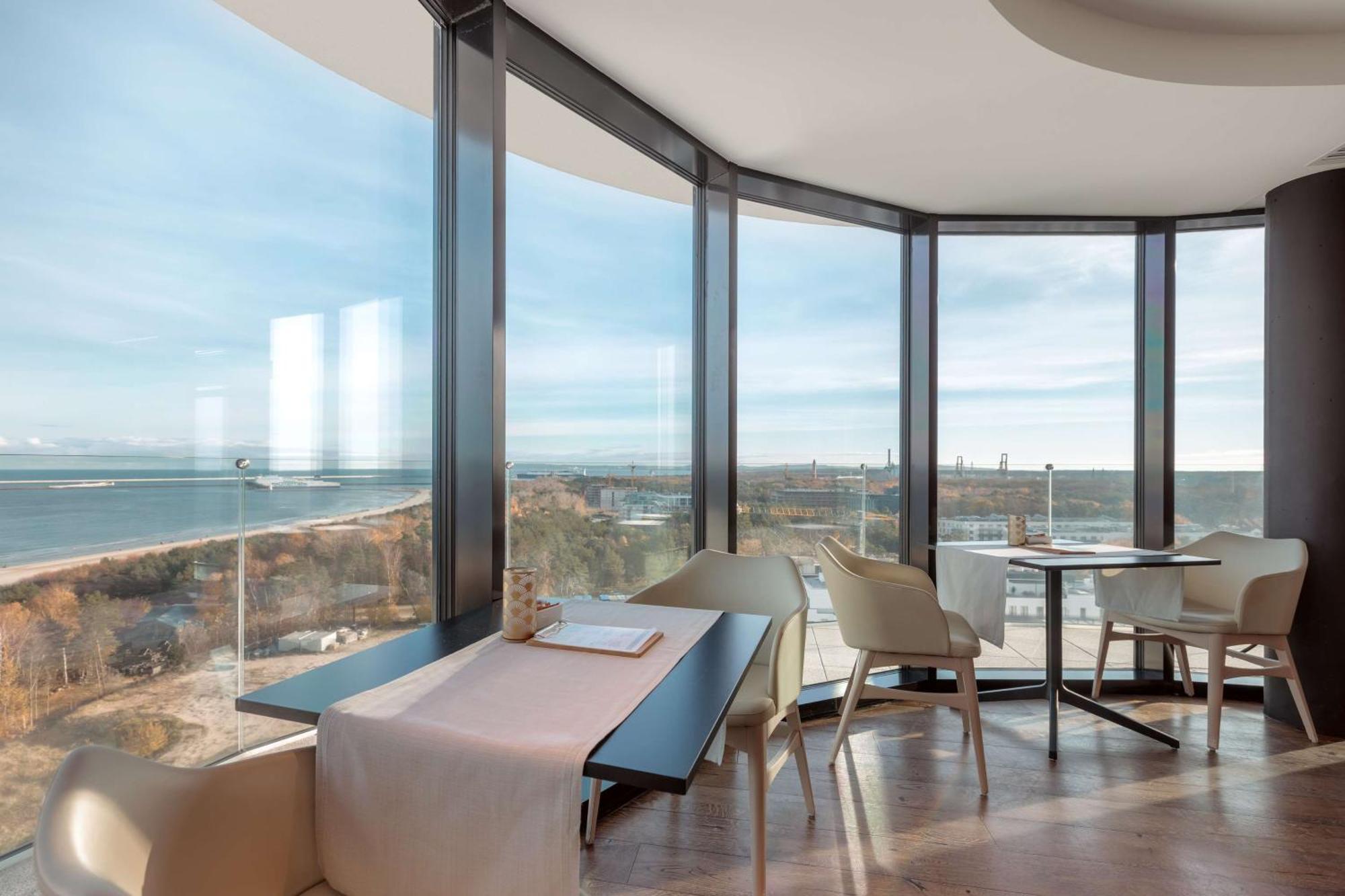 Radisson Blu Resort Świnoujście Exterior foto The photo shows a modern dining area with large windows that offer a panoramic view of the landscape outside, including a body of water and possibly a shoreline. The design features sleek furniture, including a table with chairs arranged around it, a