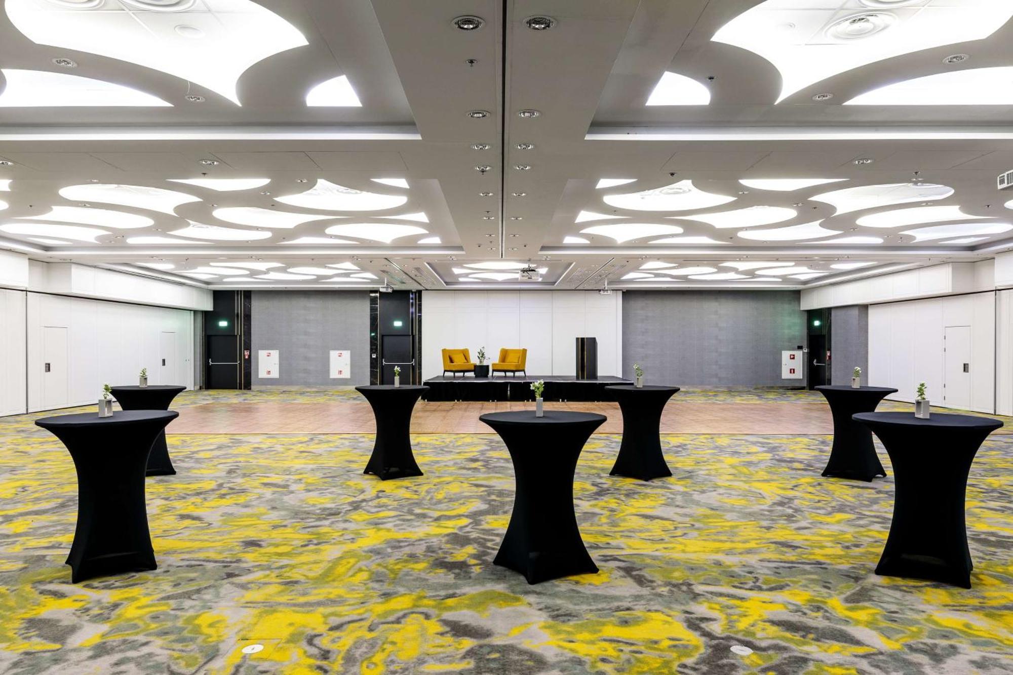 Radisson Blu Resort Świnoujście Exterior foto The photo shows a spacious interior room set up for an event. The flooring features a patterned design in gray with yellow accents. There are several black tall tables scattered throughout the area, and in the background, there are two large yellow a
