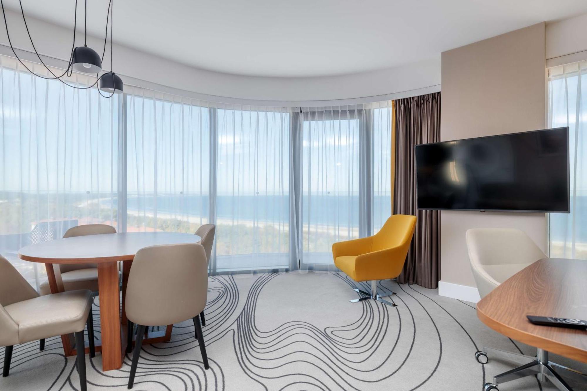 Radisson Blu Resort Świnoujście Exterior foto The photo shows a modern, stylish interior of a room with large windows that provide a panoramic view. The space features a round dining table surrounded by chairs, including a distinct yellow armchair. There is a flat-screen television mounted on th
