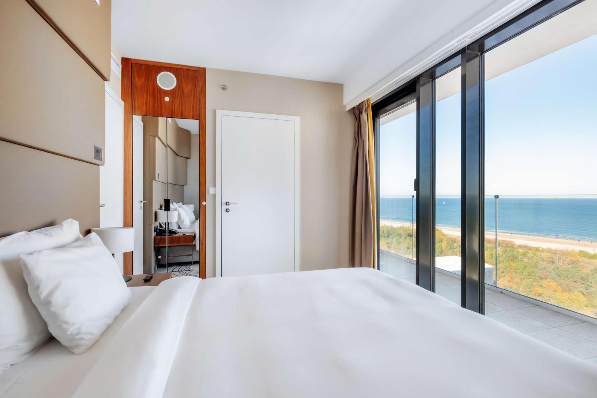 Radisson Blu Resort Świnoujście Exterior foto The photo shows a modern hotel room with a bright and airy atmosphere. There is a large bed with white bedding positioned near a glass wall that offers a view of the ocean. The room features neutral-colored walls and wooden accents, enhancing its con