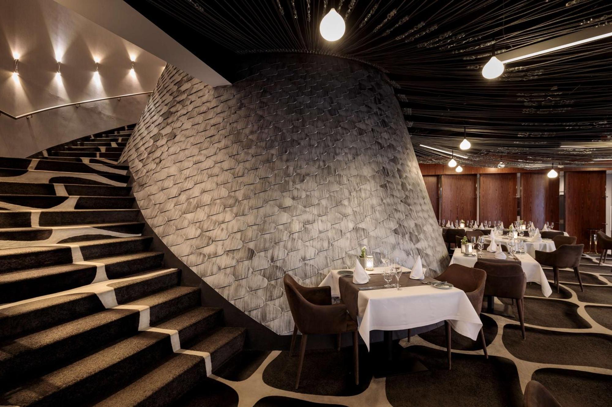 Radisson Blu Resort Świnoujście Exterior foto The photo shows the interior of a stylish restaurant. In the foreground, there are elegantly set tables with white tablecloths and dark chairs. A unique, curving stone wall adds a distinct architectural feature, and a grand staircase with a textured 