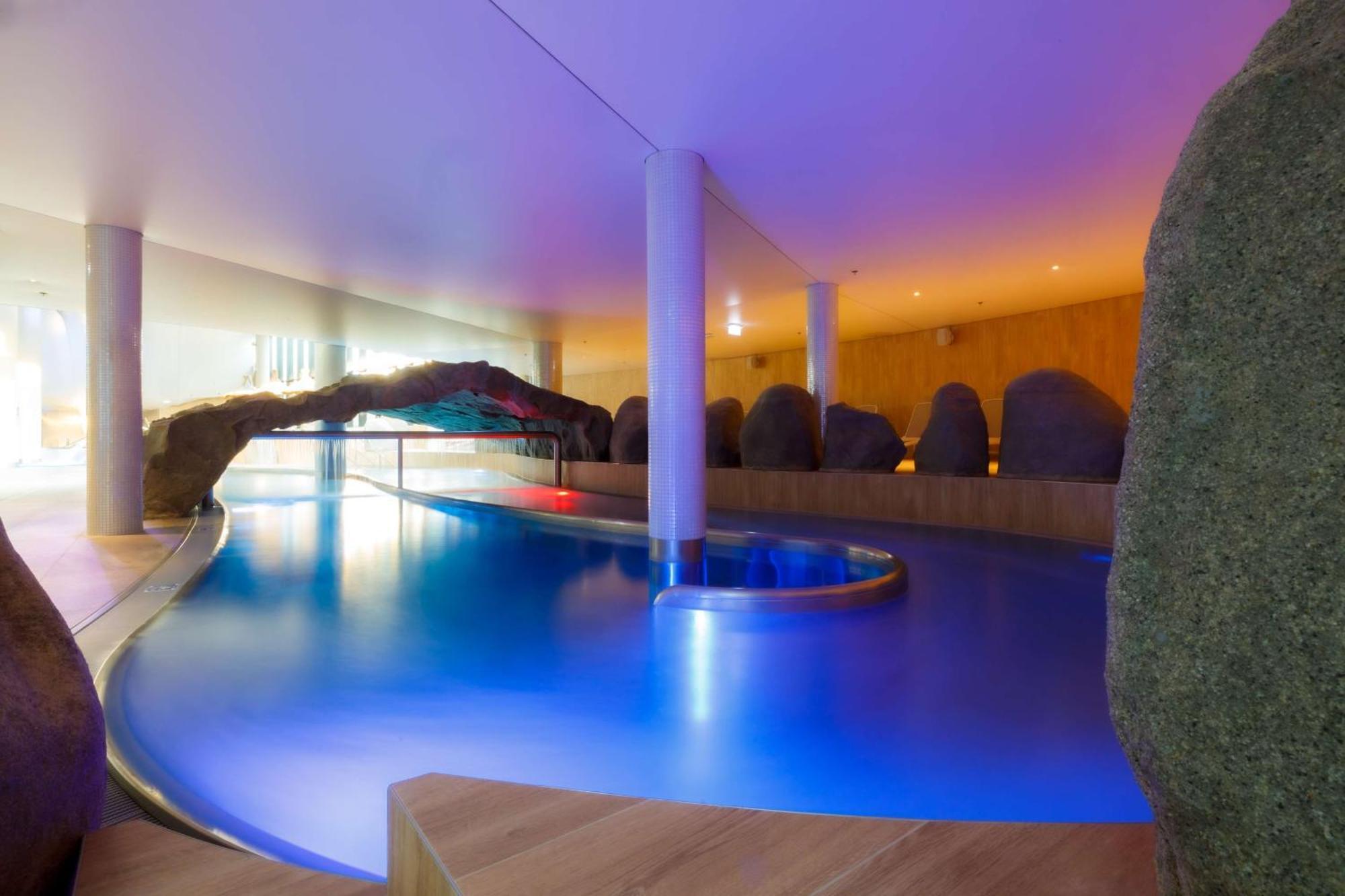 Radisson Blu Resort Świnoujście Exterior foto The photo showcases an elegant indoor swimming pool area. The pool has a flowing, organic shape and is illuminated with soft blue and purple lighting, creating a calming ambiance. Surrounding the pool are smooth, natural-looking stones and wooden ele